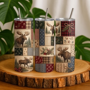 Peut inclure: Trois gobelets cylindriques en métal avec couvercles et pailles, présentant chacun un motif patchwork d'élans, de sapins, de pommes de pin et de carreaux dans des tons rouge, vert, bleu et crème. Les gobelets affichent un motif rustique.