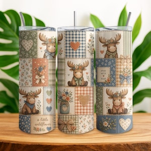 May include: A cylindrical tumbler with a straw featuring a patchwork design of moose, hearts, flowers, and bows in muted blue, green, and orange tones. The pattern includes text that reads a little wildflower and stay wild kind.