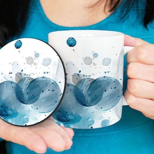 Peut inclure: Mug et dessous de verre en céramique blanche avec un motif aquarelle de cercles bleus superposés et d'éclaboussures grises et bleues. La tasse a une anse et le dessous de verre est rond.