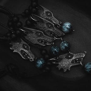 May include: Close-up of several silver-toned wolf head pendants with black cords and dark blue beads. The pendants are detailed with open mouths and sharp teeth. The background is a dark, textured surface.