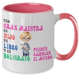 May include: White ceramic coffee mug with a pink rim and handle. The mug features a cartoon illustration of a teacher holding a book and the text "UNA GRAN MAESTRA UN HIJO UN LIBRO Y UN BOLÍGRAFO PUEDEN CAMBIAR EL MUNDO".