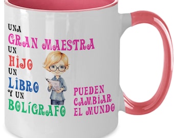 May include: White ceramic coffee mug with a pink rim and handle. The mug features a cartoon illustration of a teacher holding a book and the text "UNA GRAN MAESTRA UN HIJO UN LIBRO Y UN BOLÍGRAFO PUEDEN CAMBIAR EL MUNDO".