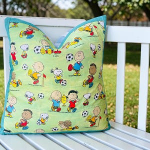 May include: Square pillow with a light green background featuring cartoon characters playing soccer. The pillow has a teal border and is resting on a white bench. The characters are in various poses with soccer balls.