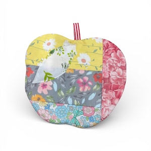 May include: A quilted apple-shaped item with a red and white striped loop. The apple is made of fabric patches in yellow, gray, pink, and blue, each with floral patterns. The item is designed with a patchwork aesthetic.