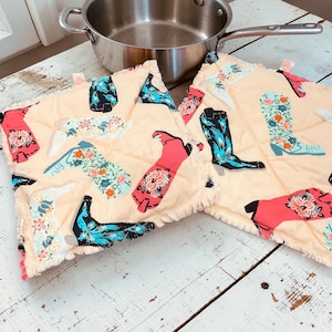 May include: Two square, quilted pot holders with a light peach background. The pot holders are decorated with colorful cowboy boot designs in red, black, and turquoise. A stainless steel saucepan with a long handle is in the background.