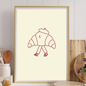 Peut inclure: Une affiche encadrée représentant un dessin au trait rouge d'un personnage croissant portant un béret et des baskets. Le croissant a un visage simple avec une petite marque en forme d'éclair et est représenté marchant sur un fond crème clair.