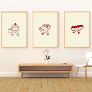 Peut inclure: Trois cadres muraux avec des fonds crème clair et des dessins au trait rouge foncé. L'impression de gauche montre un macaron avec une cerise qui marche, le centre une crêpe qui marche, et la droite un éclair qui marche, tous avec des membres.