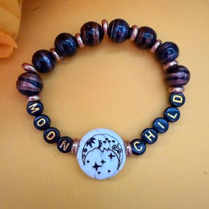 Peut inclure: Un bracelet de perles avec les mots "MOON CHILD" épelés en perles noires et dorées. Il comporte une pièce centrale circulaire blanche avec un motif céleste noir et des perles marron foncé et cuivrées.