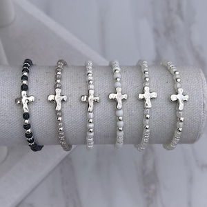 May include: Six beaded bracelets with silver cross charms. The bracelets feature black, gray, and white beads. The crosses have a hammered metal appearance. The bracelets are displayed on a white fabric roll.