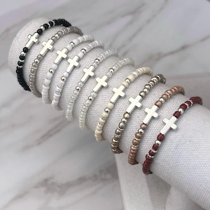May include: A collection of beaded bracelets in various colors, each featuring a silver cross charm. The bracelets are displayed on a white fabric roll, showcasing the different bead colors, including black, white, and shades of brown.