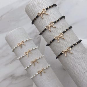 May include: Several beaded bracelets with gold-colored bow charms are displayed. Some bracelets are black with gold beads, while others are white with gold beads. The bracelets are arranged on cylindrical forms.