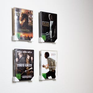 Könnte beinhalten: Vier DVD-Hüllen an einer weißen Wand, gehalten von weißen Kunststoffhaltern. Die Filme sind "Gefährten", "Gran Torino", "True Grit" und "12 Years a Slave". Jede Hülle hat ein Bewertungssymbol.
