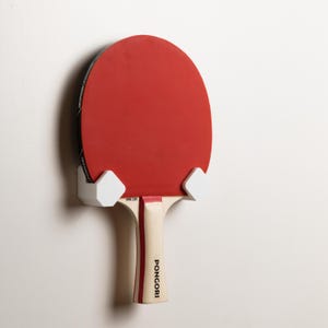 Könnte beinhalten: Eine weiße Wandhalterung mit einem rot-schwarzen Tischtennis Schläger befestigt. Der Schläger hat den Schriftzug "PONGORI" auf dem Griff.