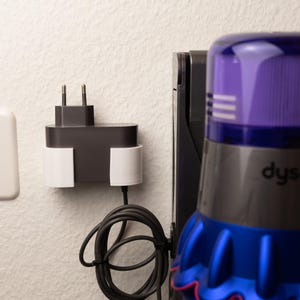 Pode incluir: Um carregador de parede cinza e preto com um cabo branco, e um aspirador de pó sem fio cinza e azul com a palavra "Dyson" nele.