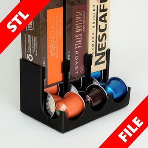 Può includere: Porta capsule di caffè in plastica nera con sei slot per capsule di caffè. Il porta capsule ha tre scatole di capsule di caffè sul retro, una con l'etichetta "Starbucks Italian Style Roast", una con l'etichetta "Starbucks Colombia" e una con l'etichetta "Nescafe Farmers Origins".