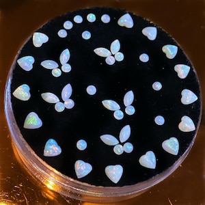 May include: Assortment of white opal cabochons in various shapes, including hearts and butterflies, displayed on a black background.
