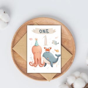 Könnte beinhalten: Geburtstagskarte mit einem Oktopus mit Partyhut, einer Krabbe und einem Wal. Die Karte zeigt das Wort "ONE" in einem Banner und die Zahl "1". Die Karte liegt auf einem Holztablett mit einem braunen Umschlag.