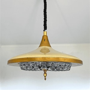 May include: A pendant light fixture featuring a wide, cream-colored metal shade with a gold-toned rim. The center includes a textured, clear glass diffuser with a diamond-cut pattern, suspended by a black cord with a gold-toned finial at the base.