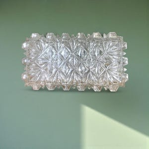 May include: A rectangular, clear glass light fixture with a geometric starburst pattern. The fixture is mounted on a light green wall. The glass has a textured, faceted surface, creating a play of light and shadow.