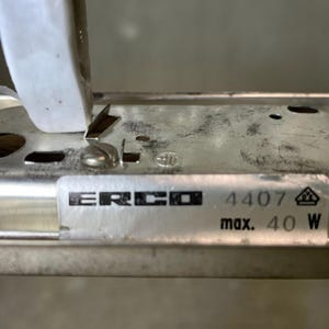 May include: Close-up of a silver metal light fitting component with the brand name "ERCO" and the numbers "4407" and "max. 40 W" printed on a label. The fitting has a screw and a small metal tab.