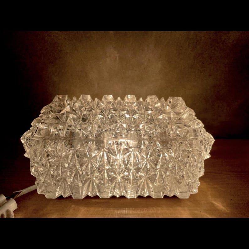 May include: A clear, rectangular glass box with a faceted, geometric design. The box has a lid and is lit from within, creating a sparkling effect. The box is on a wooden surface.