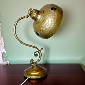 May include: An antique brass desk lamp with an ornate design. The lamp features a curved arm and a domed shade with floral engravings and three dark blue accents. The base is round and detailed, with a decorative edge. The lamp is on a dark wooden surface.
