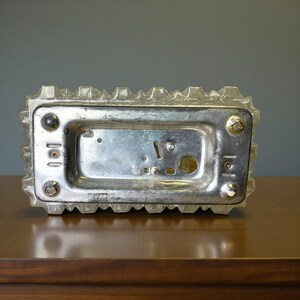 May include: A rectangular, clear glass object with a textured, ridged border and a silver metal base. The base has several screws and a central rectangular indentation. The object is resting on a wooden surface.