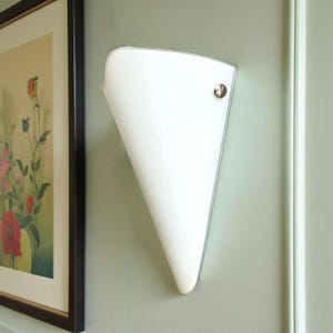 May include: A white, cone-shaped wall sconce with a brass-colored accent. The sconce is mounted on a light green wall. A framed floral print is visible in the background.
