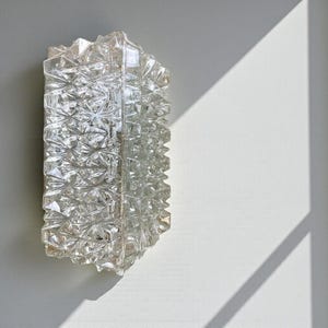 May include: A clear, rectangular wall sconce with a textured, geometric design. The sconce is made of glass and is mounted on a white wall. The light fixture has a faceted surface that reflects light.