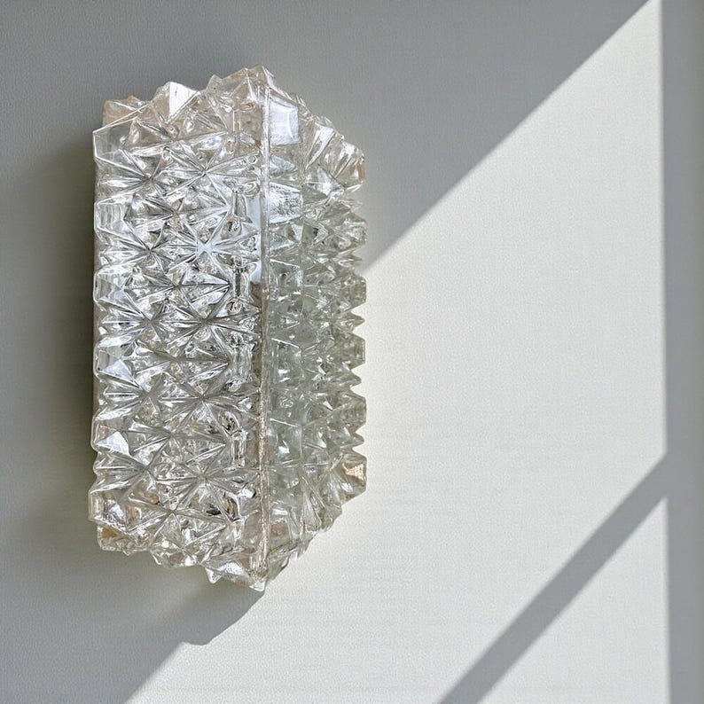 May include: A clear, rectangular wall sconce with a textured, geometric design. The sconce is made of glass and is mounted on a white wall. The light fixture has a faceted surface that reflects light.