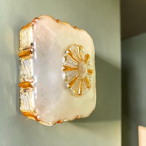 May include: A decorative wall sconce with a rectangular, textured, off-white glass base and a central, oval-shaped, amber-coloured glass design. The edges of the sconce are trimmed with amber-coloured glass.