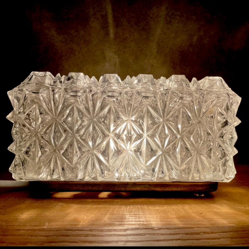 May include: A clear, rectangular glass lamp with a geometric starburst pattern. The lamp sits on a rectangular wooden base. The lamp's edges have a spiky, textured design. The background is dark, highlighting the lamp's intricate details.