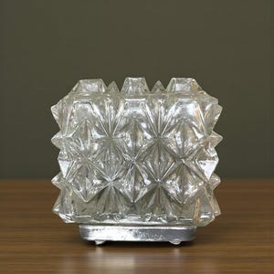 May include: A clear glass candle holder with a geometric, starburst pattern. The candle holder sits on a silver-coloured metal base. The glass has a faceted design, creating a sparkling effect.