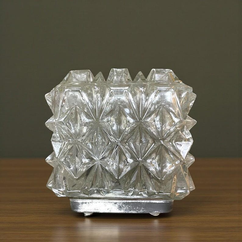 May include: A clear glass candle holder with a geometric, starburst pattern. The candle holder sits on a silver-coloured metal base. The glass has a faceted design, creating a sparkling effect.