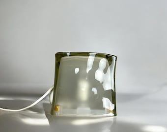 May include: A clear glass table lamp with a rounded, rectangular base. The lamp has a white cord and a light bulb inside.