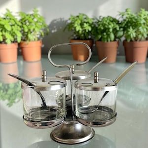 Peut inclure: Un ensemble de service à condiments composé de trois bocaux en verre transparent avec des couvercles en métal argenté et des cuillères assorties. Les bocaux sont maintenus dans un support en métal à étages avec une poignée centrale.