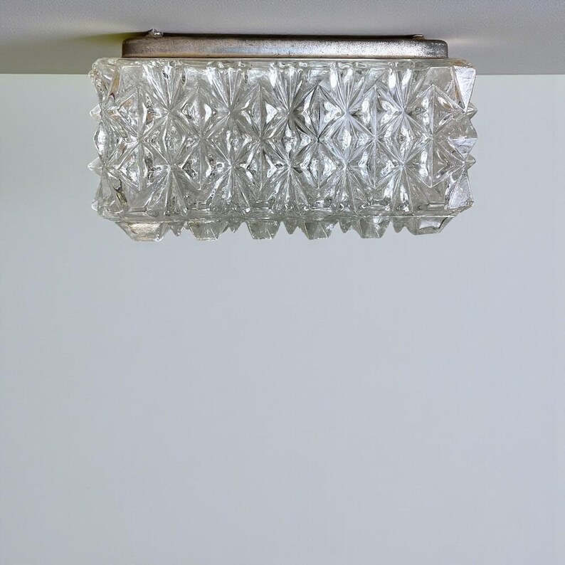 May include: A vintage, rectangular ceiling light fitting. The clear glass shade has a geometric, diamond-like pattern. The fitting has a silver-coloured metal frame. The light is mounted on a white ceiling.