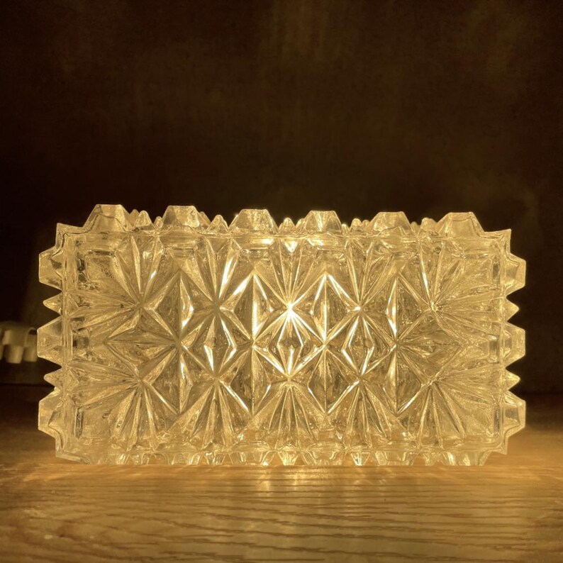 May include: A rectangular, clear glass ashtray with a geometric starburst pattern. The ashtray has a textured surface and a jagged edge. The ashtray is illuminated from within, casting a warm glow.