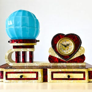 Pode incluir: Um relógio em estilo Art Déco com um globo azul claro. O relógio tem um mostrador em forma de coração e duas gavetas pequenas. O relógio tem um esquema de cores vermelho e creme. As medidas são em centímetros.