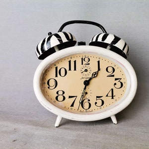 May include: A white alarm clock with black and white striped bells. The clock face is cream colored with black numbers and hands. The clock is set to 10:55.