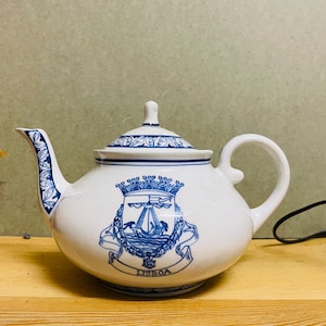 May include: White ceramic teapot with blue floral accents and a blue crest design. The crest features a ship and the word "LISBOA". The teapot has a curved spout and handle, and a lid with a small knob.