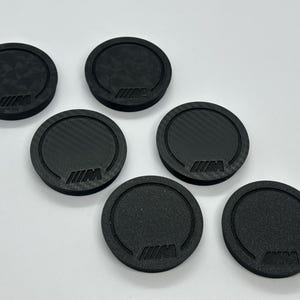 May include: Six black, round coasters with a carbon fiber texture. Each coaster features the BMW M logo in the center. The coasters are arranged on a white surface.