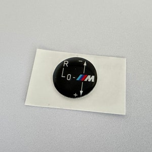 May include: A black round sticker with a white "R", "L0-", and "M" with a blue and red M logo. The sticker has a white plus and minus sign with an arrow pointing up and down.