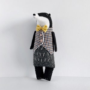 以下が含まれることがあります： 黒と白のフェルトで作られたアナグマのぬいぐるみ。茶色と白の格子柄のベストを着て、黄色の蝶ネクタイをしています。
