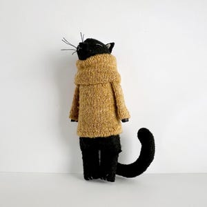 Op de afbeelding: Een handgemaakte zwarte kattenpop met een bruin gebreide trui. De kat heeft een zwarte kop met snorharen en een gebogen zwarte staart. De pop staat tegen een witte achtergrond.