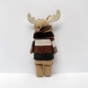 Puede incluir: Un juguete de peluche con forma de alce con cuerpo marrón, patas color beige y grandes astas color crema. El alce lleva un suéter a rayas en tonos marrones, grises y crema, y una bufanda marrón. El juguete está sobre un fondo blanco.