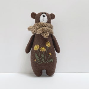 Peut inclure: Une peluche artisanale en forme d'ours brun, avec un museau blanc et une écharpe marron. L'ours porte des broderies de fleurs de pissenlit jaunes sur sa poitrine. Le jouet est en tissu doux et convient aux enfants.