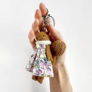 Puede incluir: Un llavero de ardilla marrón hecho a mano con una bufanda blanca de punto y un vestido floral en tonos morados, azules y blancos. La cola de la ardilla es una forma redonda y marrón con costuras blancas. El llavero se sujeta con un cierre plateado.