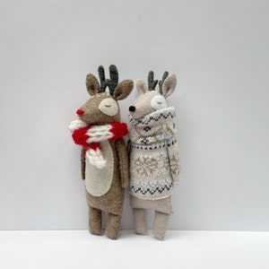 May include: Two handmade reindeer dolls. One is brown with a red and white scarf, the other is beige and wearing a patterned sweater. Both have antlers and closed eyes, suggesting a peaceful, festive theme.