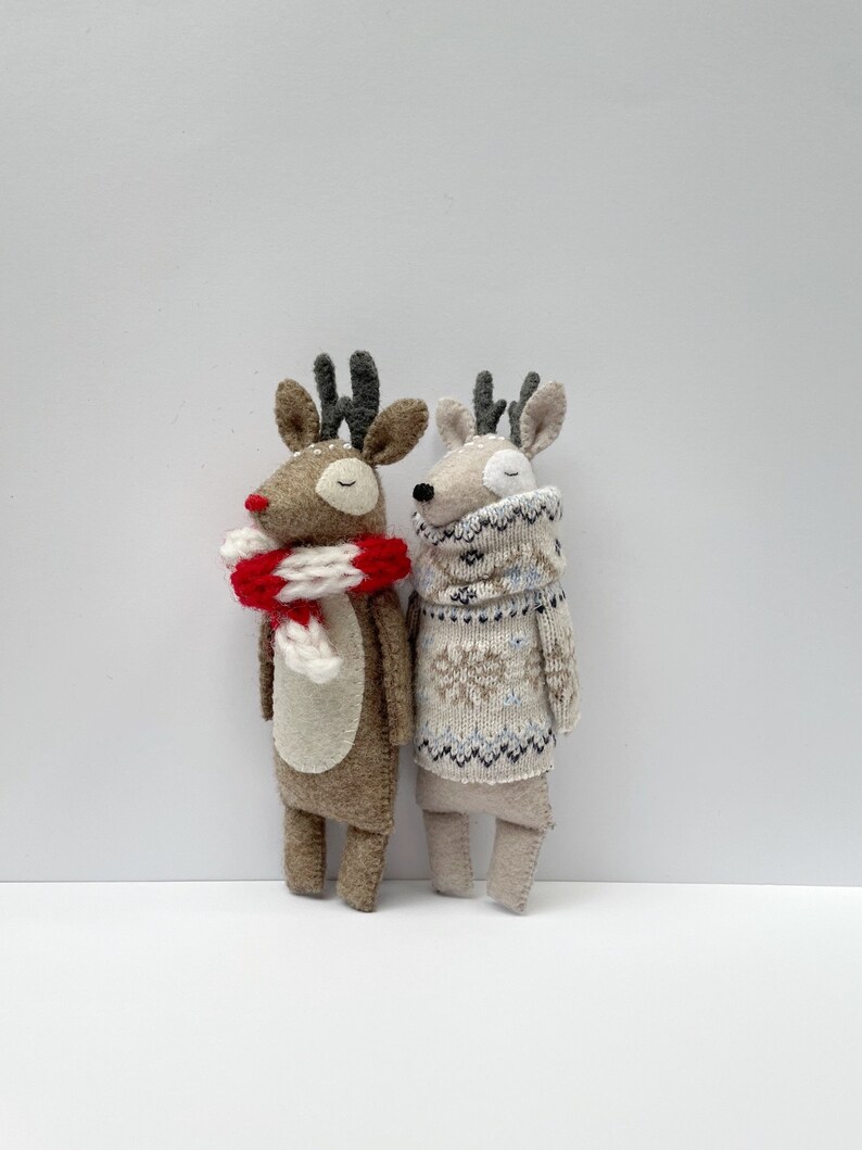 May include: Two handmade reindeer dolls. One is brown with a red and white scarf, the other is beige and wearing a patterned sweater. Both have antlers and closed eyes, suggesting a peaceful, festive theme.